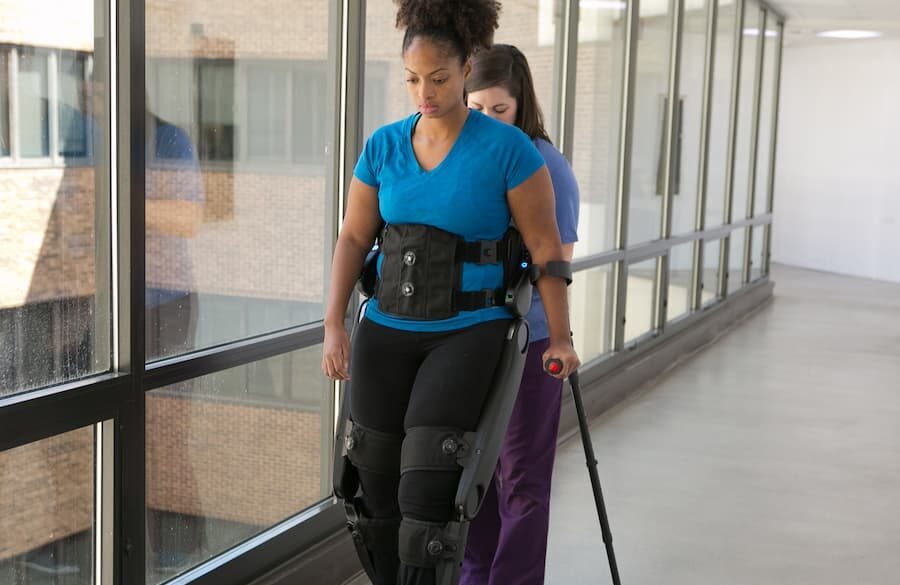 A woman in a blue shirt uses a robotic exoskeleton and a cane to walk in a hallway, assisted by another person standing behind her. Large windows line one side of the hallway.