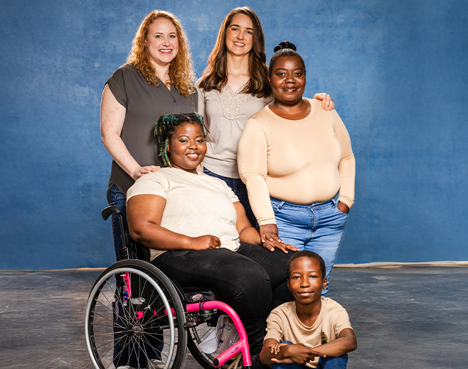 Five people of diverse backgrounds pose together, smiling. One woman sits in a wheelchair with pink accents. Two women stand behind, and another woman and a young child kneel and sit in front, all against a blue studio backdrop.