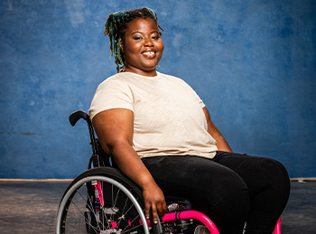 A smiling woman with braids sits in a wheelchair with pink accents against a blue textured background. She is wearing a beige t-shirt and black pants.