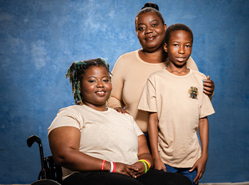 Three people pose for a portrait against a blue background. A woman in a wheelchair sits in front, while another woman and a young boy stand behind her. All three wear light-colored shirts and smile gently.
