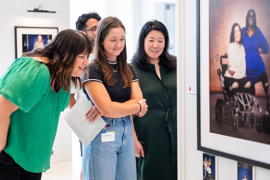 Three women and one man smile and look closely at a framed photograph on the wall in an art gallery. The atmosphere is bright and engaging, with other photos visible in the background.
