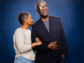 A woman with short curly hair leans gently on the arm of a man wearing glasses and a dark blazer. They both have serious expressions and stand together against a dark blue background.