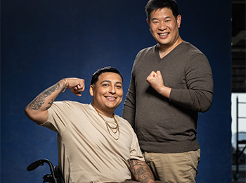 Two men smiling at the camera; one sits in a wheelchair flexing his arm, while the other stands beside him making a fist with a supportive gesture. Both appear happy and confident against a dark blue background.