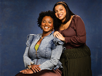 Two women pose together in front of a blue background. One woman is seated, smiling, wearing a denim jacket and skirt. The other stands beside her, smiling, dressed in a brown blouse and dark skirt, with her hand on the seated woman's shoulder.