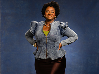 A woman stands confidently against a blue backdrop, wearing a ruffled denim jacket over a green top and a dark skirt. She has short curly hair, hands on her hips, and accessorizes with jewelry.