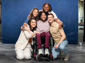 Five people pose together, smiling warmly. One person sits in a wheelchair at the center, while the others kneel or stand beside and behind, embracing and supporting each other against a blue backdrop.