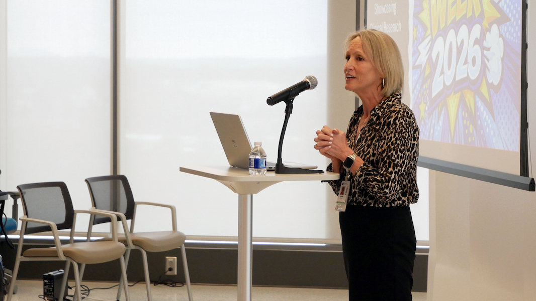A woman stands at a podium with a laptop and microphone, speaking to an audience. Behind her, a projection displays colorful graphics and the text “WEEK 2026.” There are empty chairs and large windows in the background.
