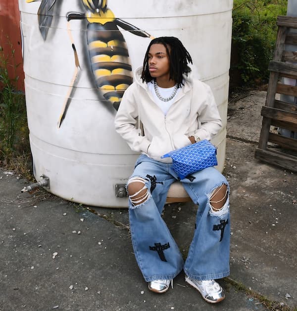 A young man with locks sits by a large white tank with a wasp mural. He wears a white hoodie, ripped jeans with black cross patches, shiny silver shoes, and holds a blue textured bag.