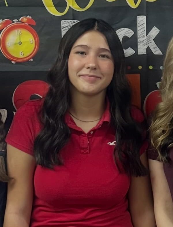 A young woman with long, wavy dark hair wearing a red collared shirt stands in front of a colorful 