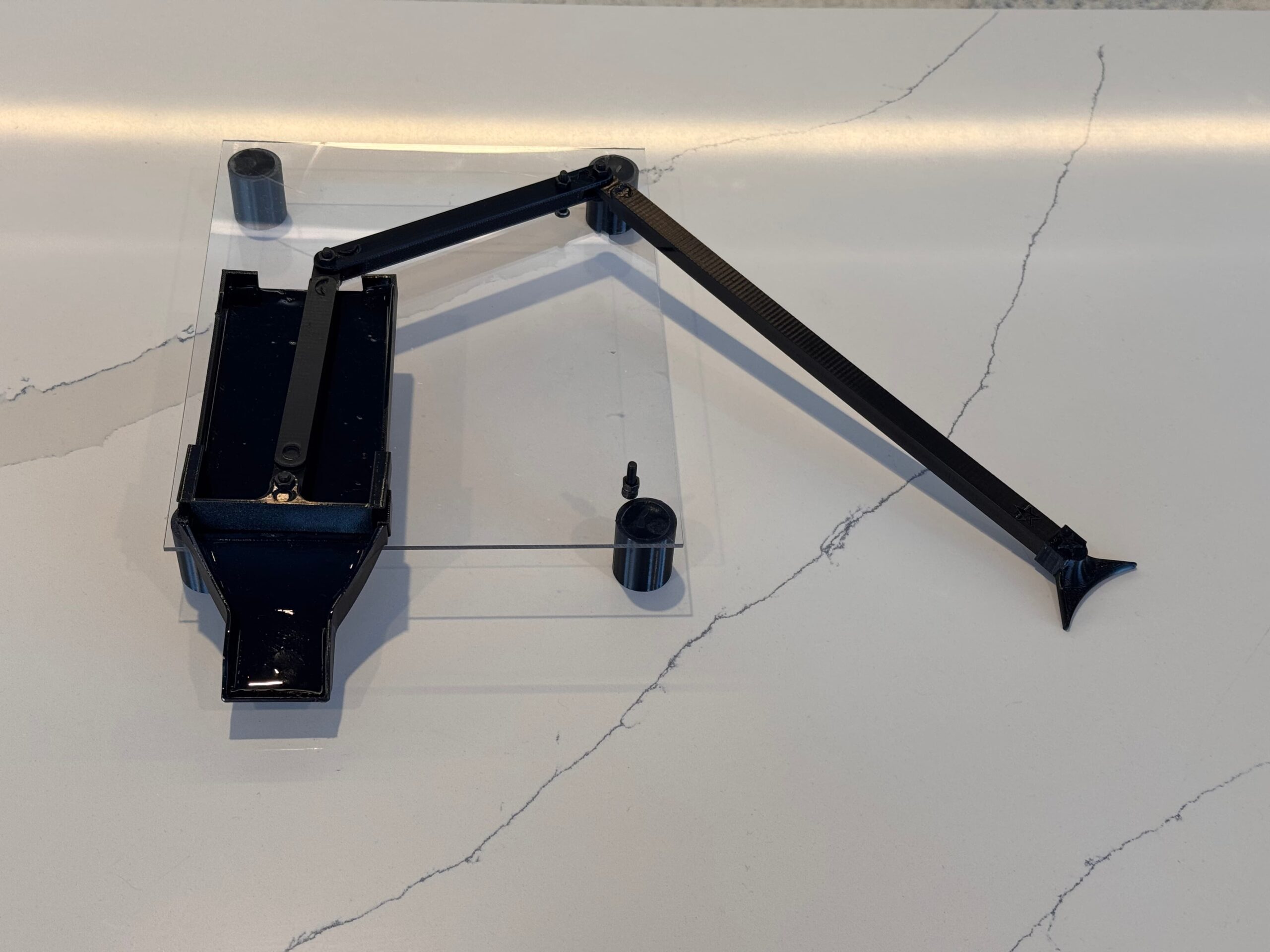 A black mechanical arm with jointed segments is mounted on a clear rectangular base, resting on a white marble surface with grey veining. A small loose screw sits near the center of the base.