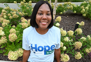 A woman smiles while sitting in a garden with green flowering plants. She wears a light blue shirt with 