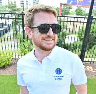 A man with red hair and a beard, wearing black sunglasses and a white Shepherd Center polo shirt, stands outside by a fence and greenery, smiling.