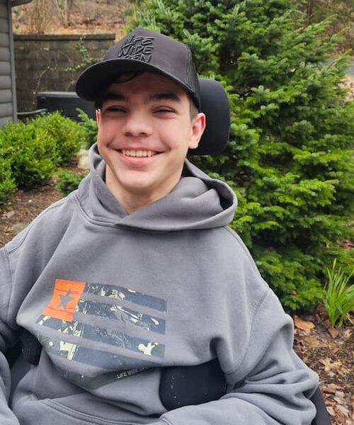 A smiling teenage boy wearing a dark cap and gray hoodie sits in a wheelchair outdoors near green bushes and a small pine tree.