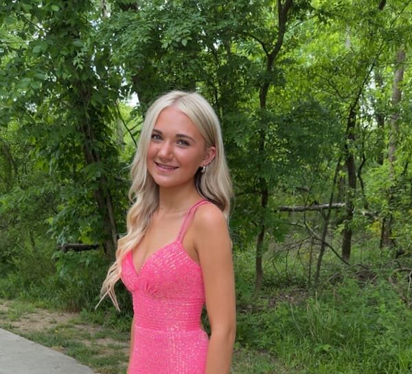 A young woman with long blonde hair wearing a bright pink dress stands smiling outdoors in front of lush green trees and foliage.